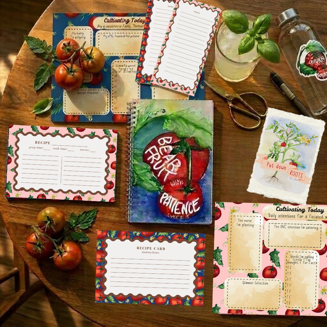 A styled flat-lay of multiple recipe cards, a spiral notebook, and a small card set arranged on a wooden table with tomatoes, basil, scissors, and a glass bottle nearby.