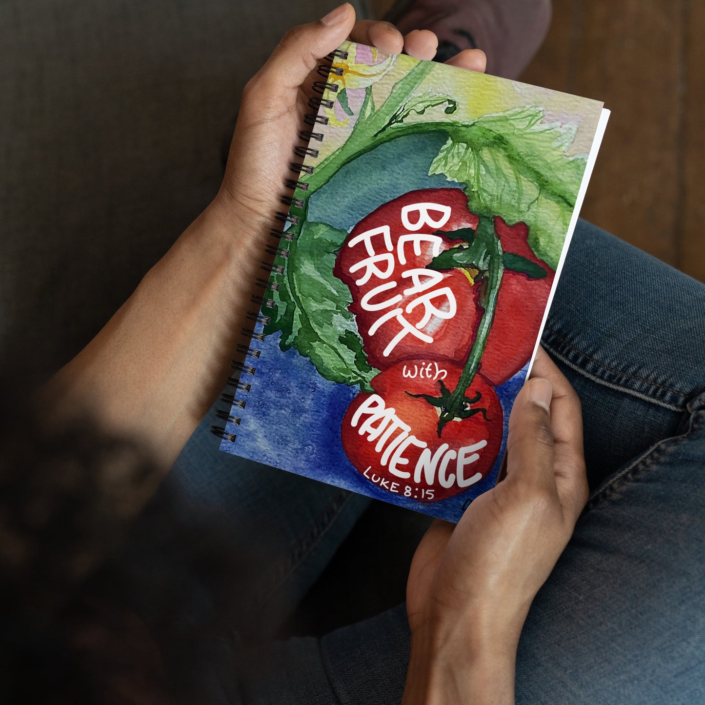 A person holding a spiral-bound notebook with a colorful watercolor illustration of red tomatoes on the cover, with the text 'BEAR FRUIT WITH PATIENCE" LUKE 8:15'.