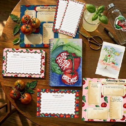 A styled flat-lay of tomato-themed recipe cards, a spiral-bound recipe book titled 'Bear Paw Patience', and other kitchen planning items arranged on a wooden table with tomatoes, basil, scissors, and a glass bottle.