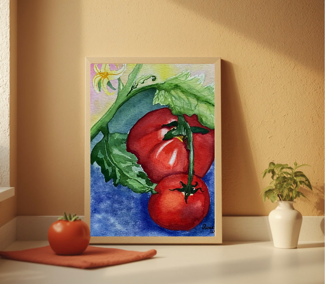 A framed watercolor painting of two red tomatoes on a vine with green leaves and a blue background, displayed on a shelf against a beige wall with a small plant and a tomato prop nearby.